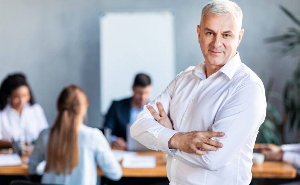 Business And Entrepreneurship. Mature Businessman At Corporate Meeting Standing Smiling To Camera In Modern Office. Free Space